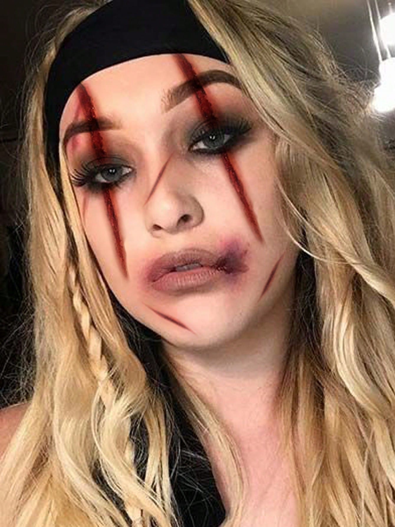 A person with wavy blonde hair and a black headband wears horror-themed makeup with gash-like lines and bruising, creating a zombie-like look.