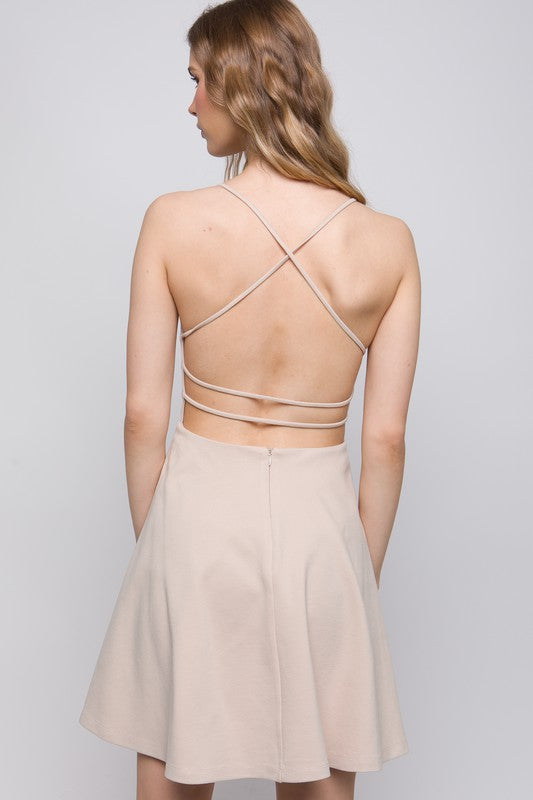 the dress is a light beige, knee-length A-line with a delicate back design featuring thin crisscross straps that wrap around the back, giving a soft, feminine silhouette.
