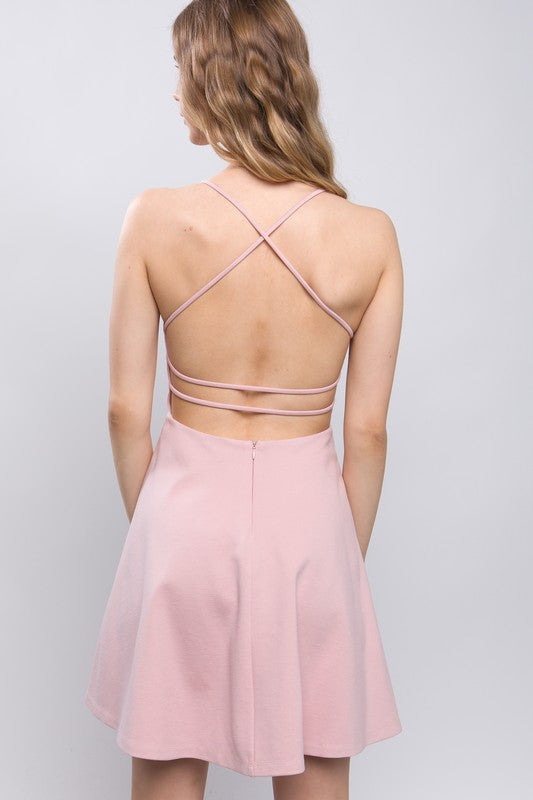 the dress is a light beige, knee-length A-line with a delicate back design featuring thin crisscross straps that wrap around the back, giving a soft, feminine silhouette.