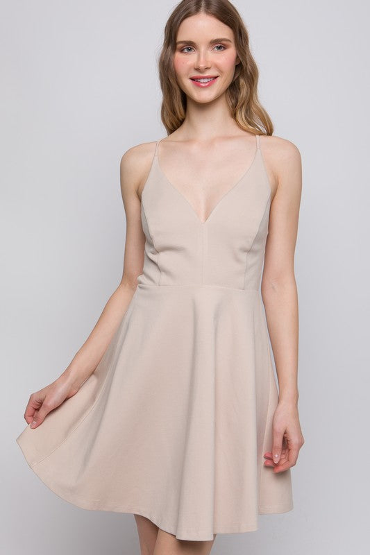 the dress is a light beige, knee-length A-line with a delicate back design featuring thin crisscross straps that wrap around the back, giving a soft, feminine silhouette.
