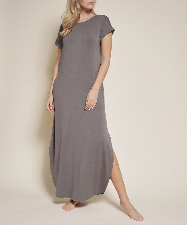 The image shows a woman wearing a comfy-looking brown maxi dress with short sleeves and a pocket on the side. She's barefoot, posing casually with one hand in her pocket and the other touching her hair against a plain beige background. Slaying the effortless vibe.