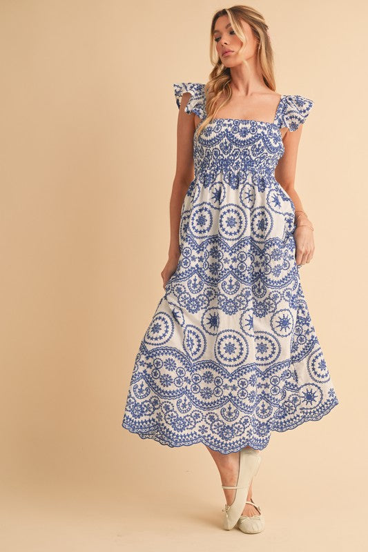 The dress is a long, airy maxi with a blue and white geometric/medallion print, cap sleeves, and a fitted bodice that flows into a relaxed, ankle-length skirt.