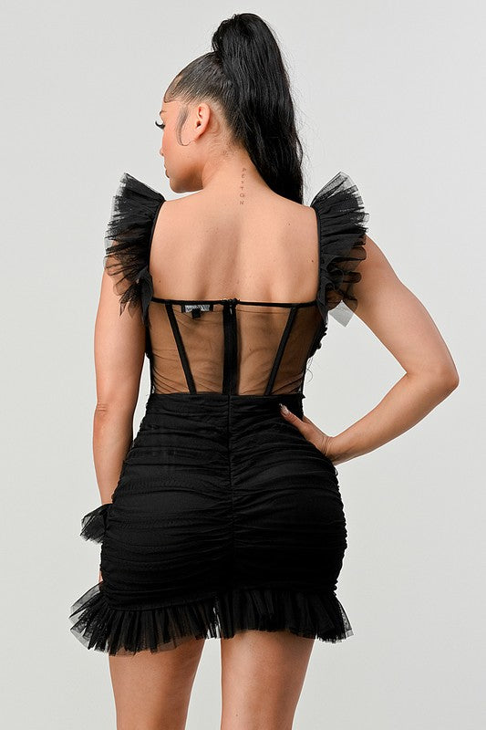 The dress is a chic black mini with a fitted silhouette, sheer paneling at the bodice, and dramatic ruffled cap sleeves that add a flirty, edgy touch