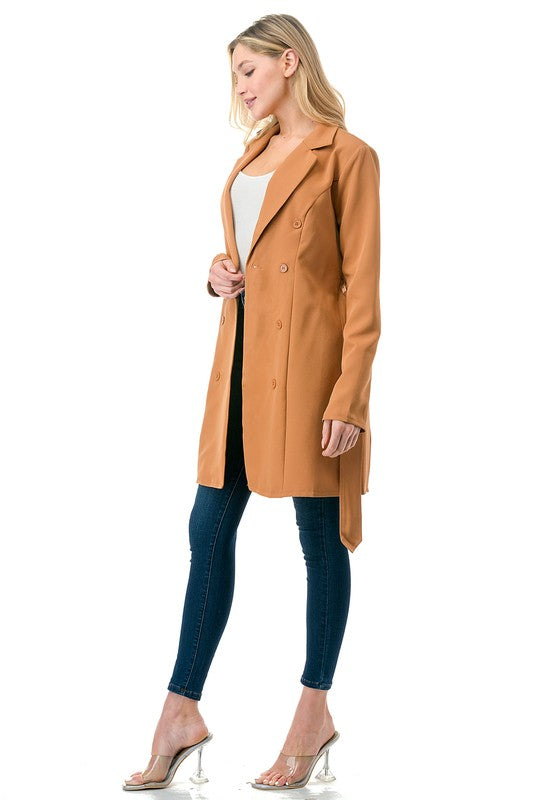 A woman wearing a long tan coat with big buttons, a white top, and blue jeans. Background's plain white with a black border.