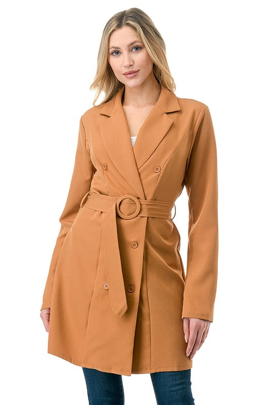 A woman wearing a long tan coat with big buttons, a white top, and blue jeans. Background's plain white with a black border.