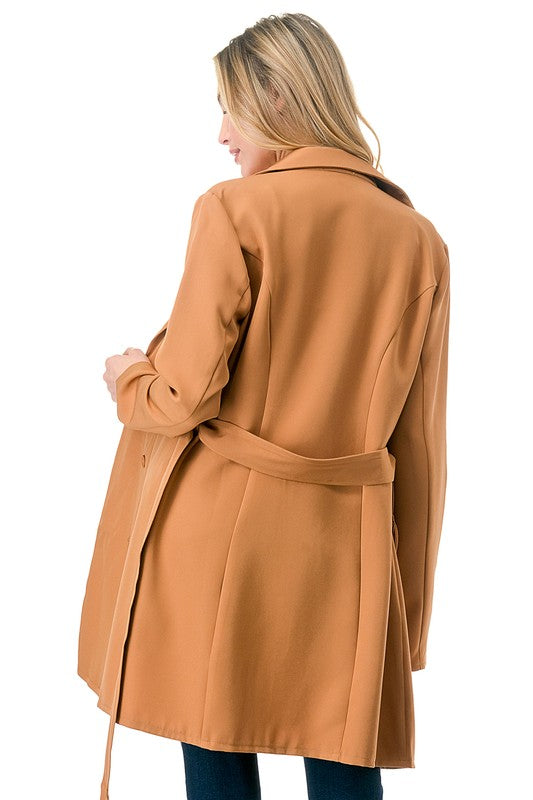 A woman wearing a long tan coat with big buttons, a white top, and blue jeans. Background's plain white with a black border.
