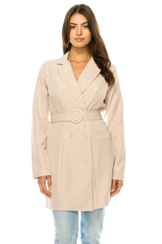 A woman wearing a long cream coat with big buttons, a white top, and blue jeans. Background's plain white with a black border.
