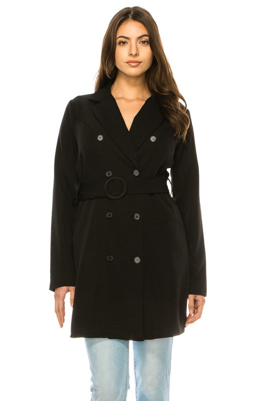 A woman wearing a long black coat with big buttons, a white top, and blue jeans. Background's plain white with a black border.
