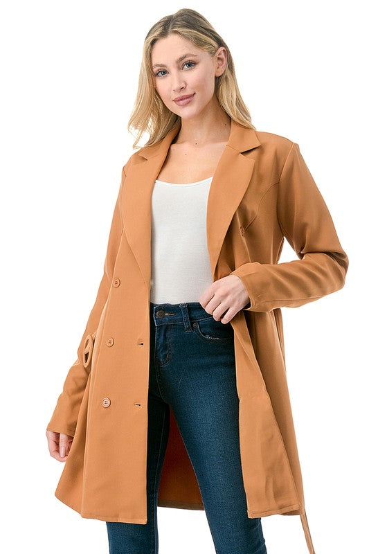 A woman wearing a long tan coat with big buttons, a white top, and blue jeans. Background's plain white with a black border.
