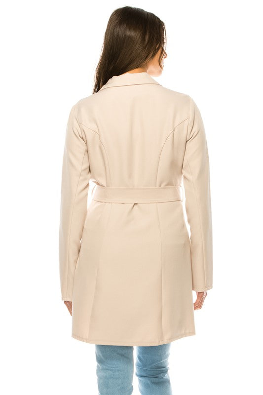 A woman wearing a long cream coat with big buttons, a white top, and blue jeans. Background's plain white with a black border.