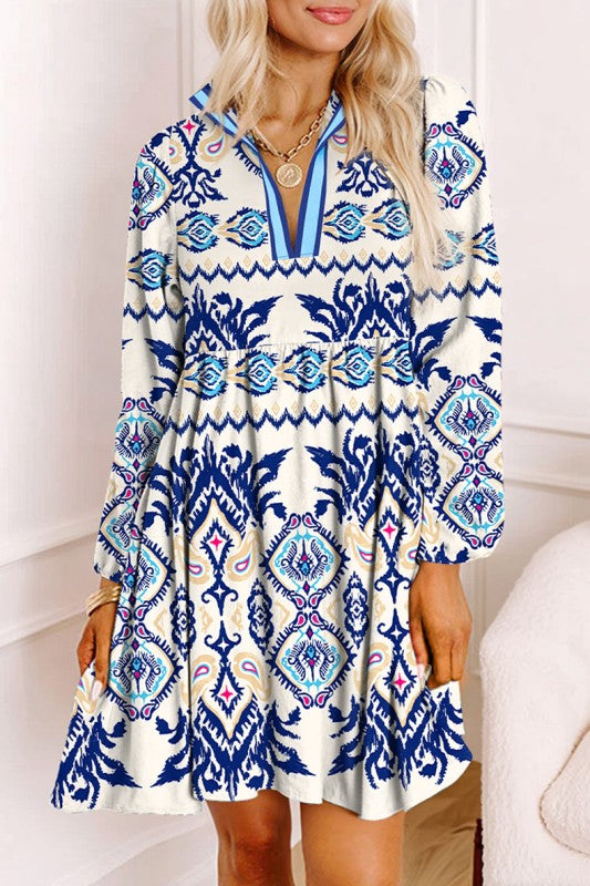 Woman wearing a blue ethnic print mini dress with striped patterns, split neckline, and long sleeves, standing indoors against a white paneled wall.