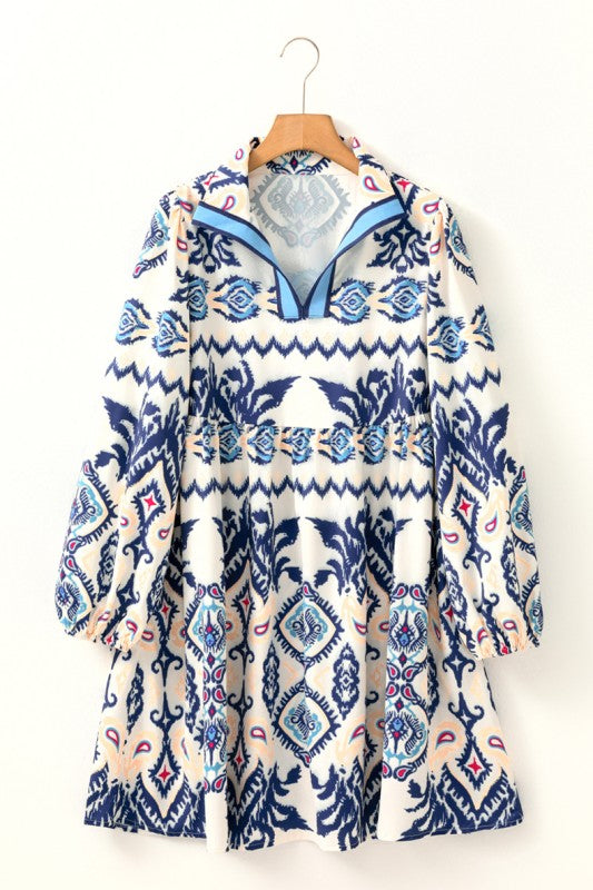 Woman wearing a blue ethnic print mini dress with striped patterns, split neckline, and long sleeves, standing indoors against a white paneled wall.