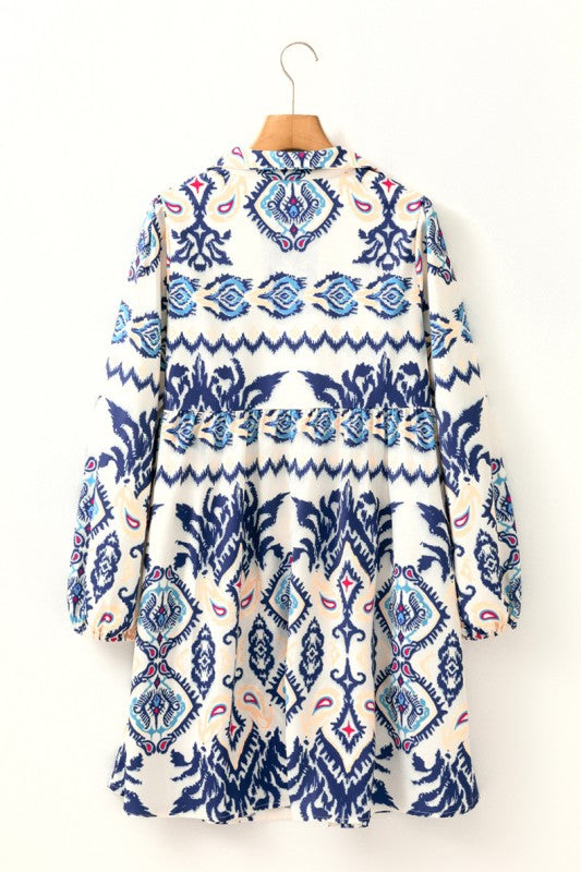 Woman wearing a blue ethnic print mini dress with striped patterns, split neckline, and long sleeves, standing indoors against a white paneled wall.