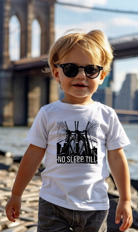 Child wearing a white t-shirt with a graphic design and text, standing in front of a bridge.