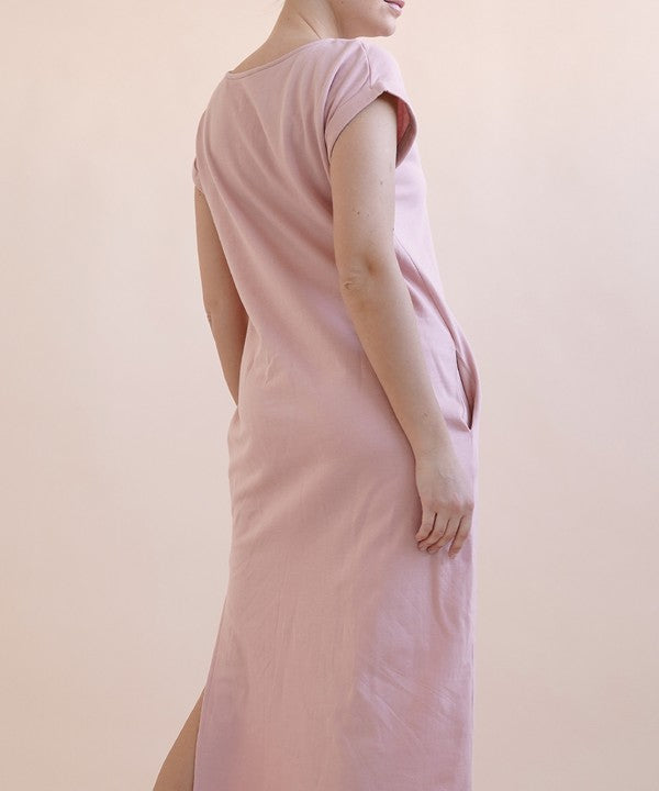 A woman wearing a light pink, short-sleeve, midi-length casual dress with side pockets, paired with black flat sandals.