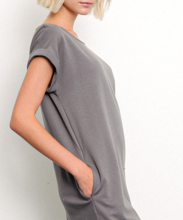 Woman in a stylish gray dress with a side slit, posing against a minimalist white backdrop. One hand is tucked into her pocket, while the other hangs relaxed by her side, with bare feet adding a touch of casual elegance.
