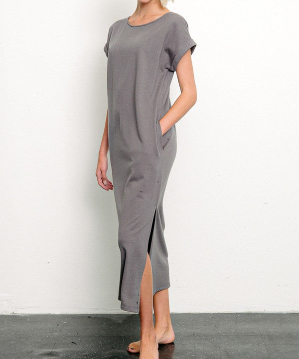 Woman in a stylish gray dress with a side slit, posing against a minimalist white backdrop. One hand is tucked into her pocket, while the other hangs relaxed by her side, with bare feet adding a touch of casual elegance.