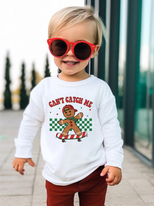 Child wearing a white sweatshirt with a gingerbread man graphic and text, standing outdoors.