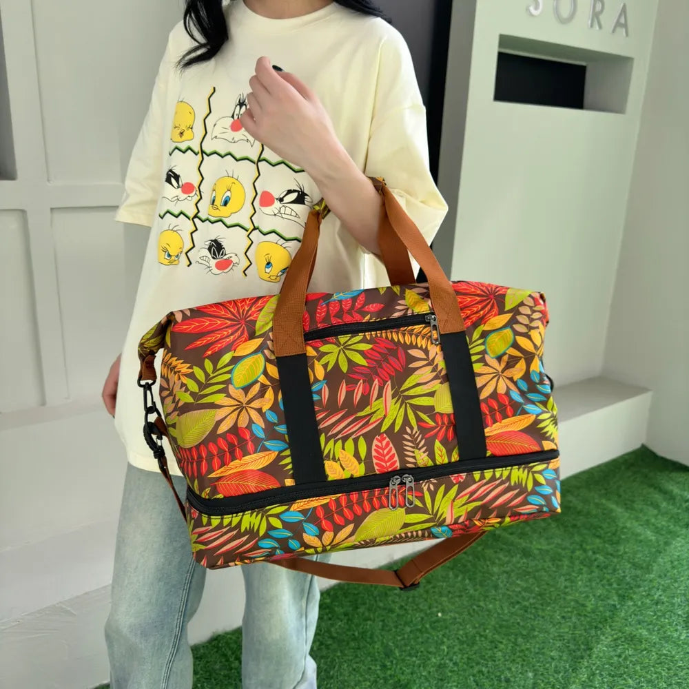 Person standing on a green carpeted floor, holding a colorful duffel bag with a vibrant leaf pattern in red, yellow, green, and blue. They're wearing a white t-shirt with cartoon duck faces and light blue jeans—fun, casual, and eye-catching.