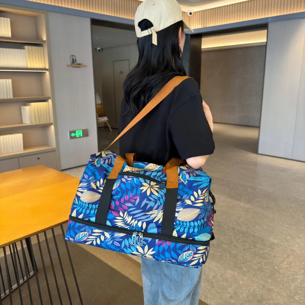 Person holding a vibrant duffel bag with blue, yellow, and green leaf patterns, wearing a black Spanish-text t-shirt and light blue jeans. Stylish and bold—perfect for casual outings or travel.