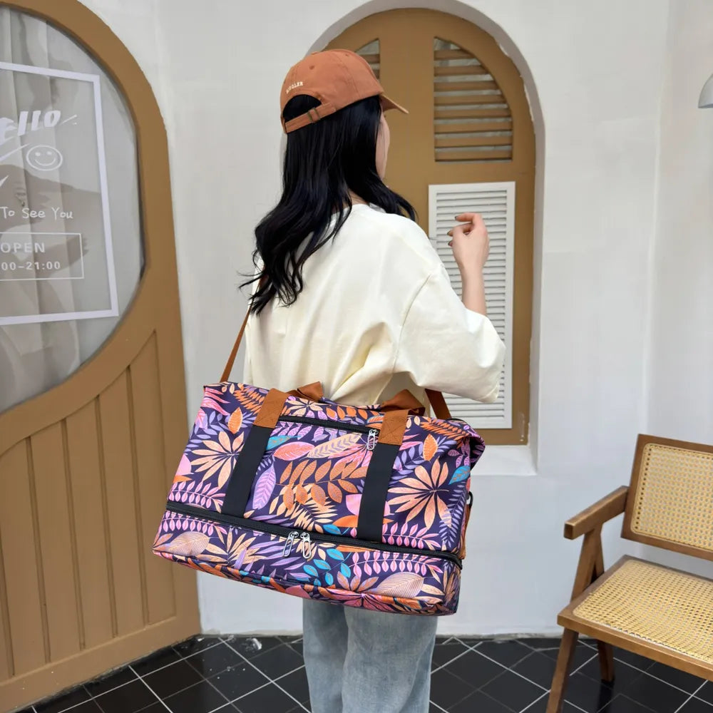 Person holding a vibrant duffel bag with a bold leaf pattern in purple, orange, pink, and yellow. They're dressed in a light yellow graphic t-shirt and light blue jeans, standing indoors near a wooden chair—fun, casual, and full of personality.