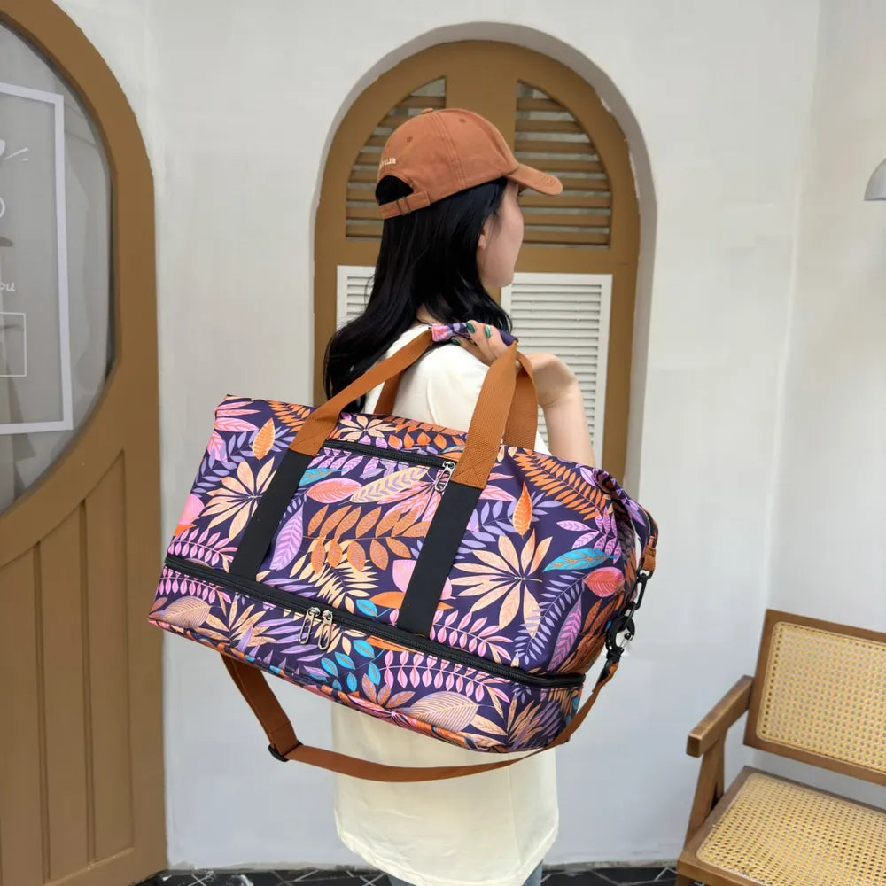 Person holding a vibrant duffel bag with a bold leaf pattern in purple, orange, pink, and yellow. They're dressed in a light yellow graphic t-shirt and light blue jeans, standing indoors near a wooden chair—fun, casual, and full of personality.