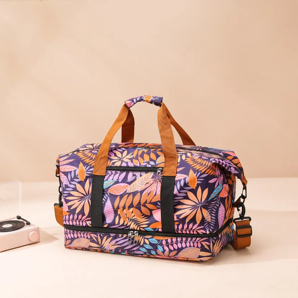 Colorful duffel bag with a vibrant leaf pattern in orange, purple, pink, and blue, featuring brown handles, a shoulder strap, and black trim. Set beside a small turntable—stylish, spacious, and perfect for travel or casual outings.