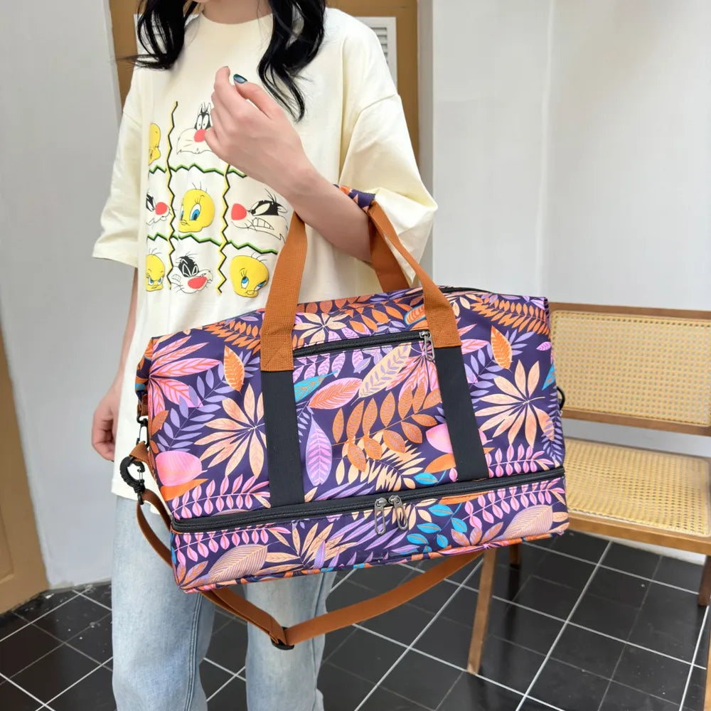 Person holding a vibrant duffel bag with a bold leaf pattern in purple, orange, pink, and yellow. They're dressed in a light yellow graphic t-shirt and light blue jeans, standing indoors near a wooden chair—fun, casual, and full of personality.