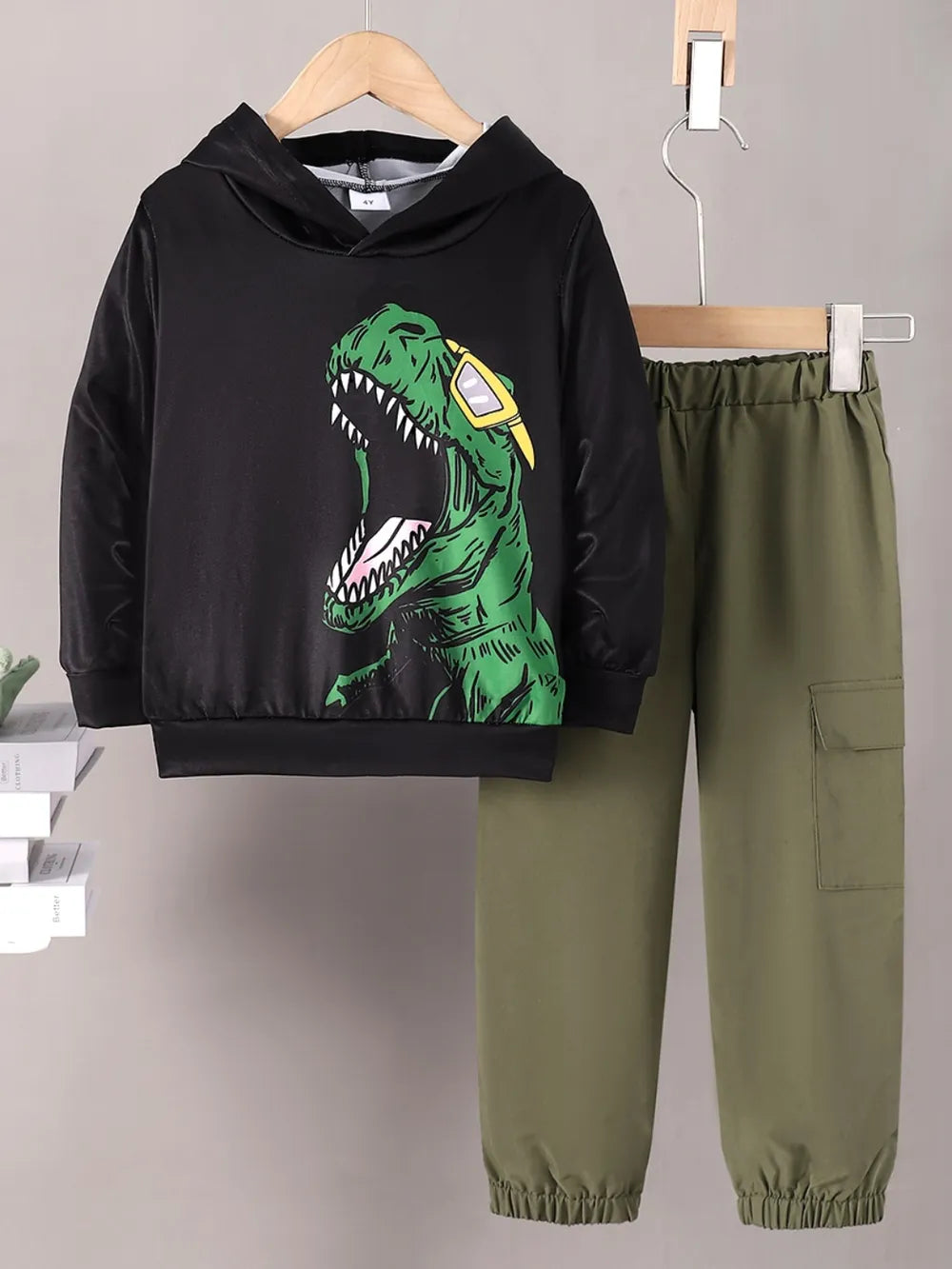 Black hoodie with green dinosaur design and green pants on hangers against a gray background