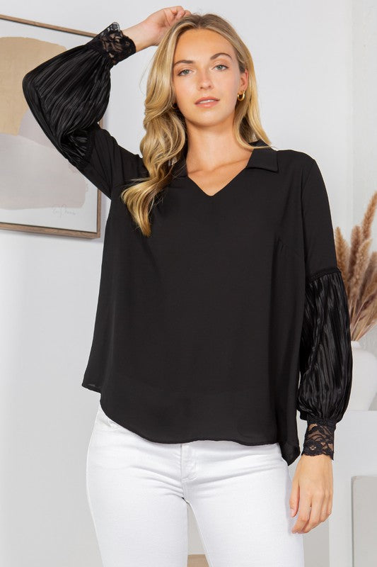 A woman with long blonde hair is posing in a black blouse with frilly sleeves and white pants. She's sitting on a wooden stool with one hand on her hip. There's a white wall behind her with a framed picture and some dried pampas grass in a vase.