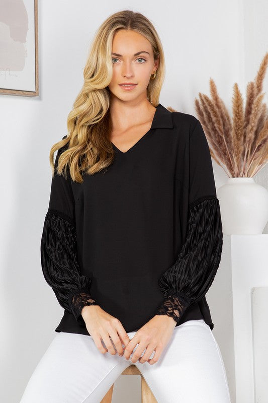A woman with long blonde hair is posing in a black blouse with frilly sleeves and white pants. She's sitting on a wooden stool with one hand on her hip. There's a white wall behind her with a framed picture and some dried pampas grass in a vase.