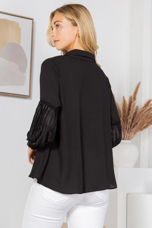 A woman with long blonde hair is posing in a black blouse with frilly sleeves and white pants. She's sitting on a wooden stool with one hand on her hip. There's a white wall behind her with a framed picture and some dried pampas grass in a vase.