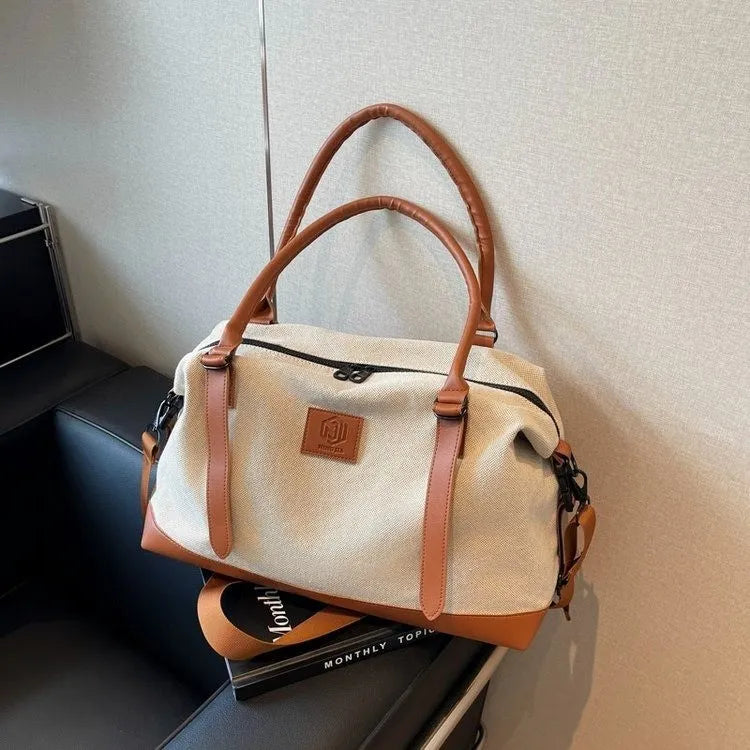 Beige and brown handbag with leather handles, shoulder strap, and embossed logo patch—placed on a black couch over a book titled "MONTHLY TOPIC."