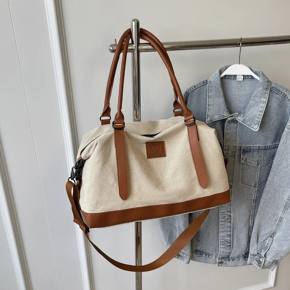 Beige and brown handbag with leather handles, shoulder strap, and vertical accents—hanging beside a denim jacket on a metal rack against a white paneled wall.