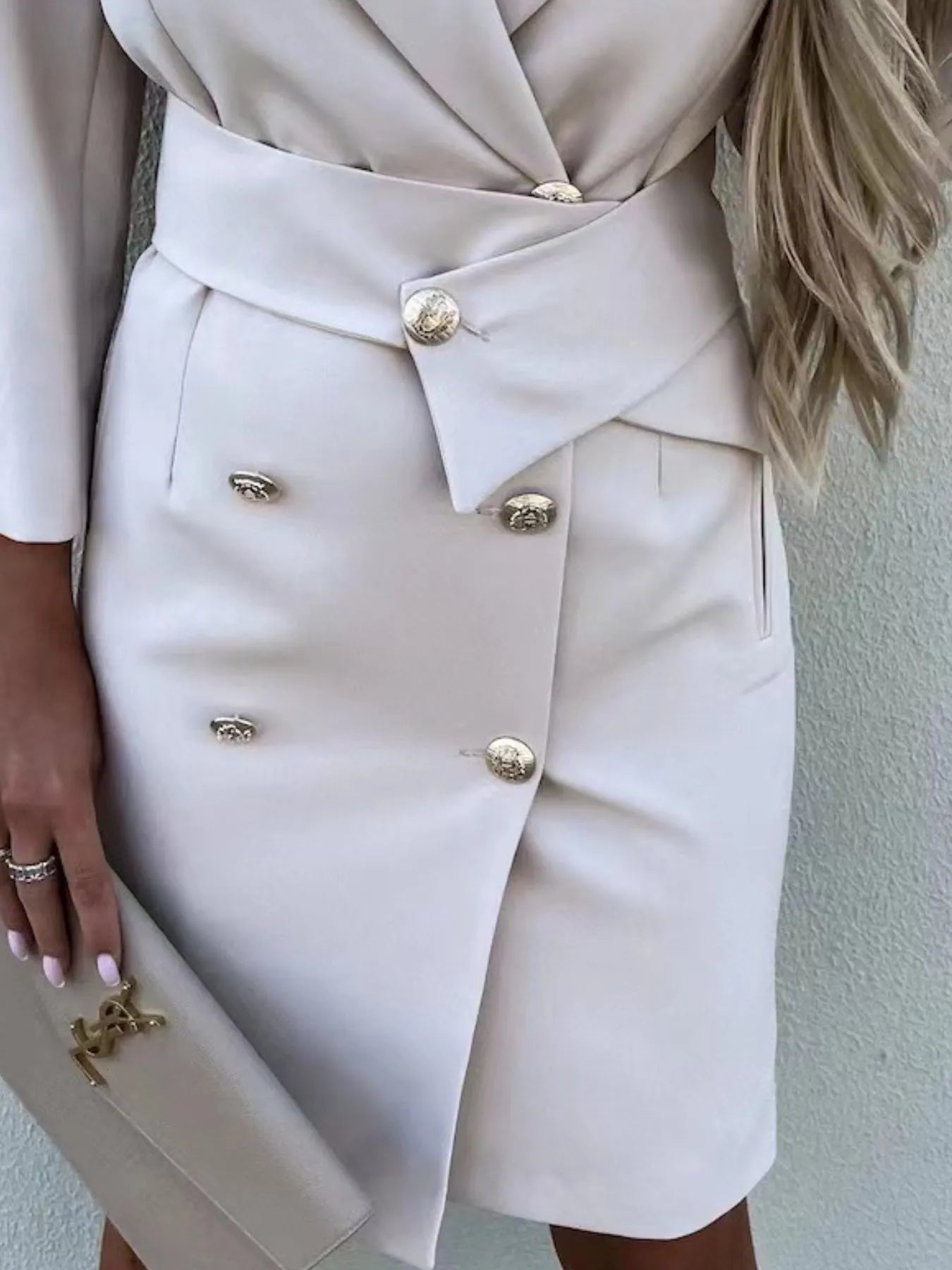 A woman in a stylish white double-breasted blazer dress with gold buttons, accessorized with jewelry and a clutch.