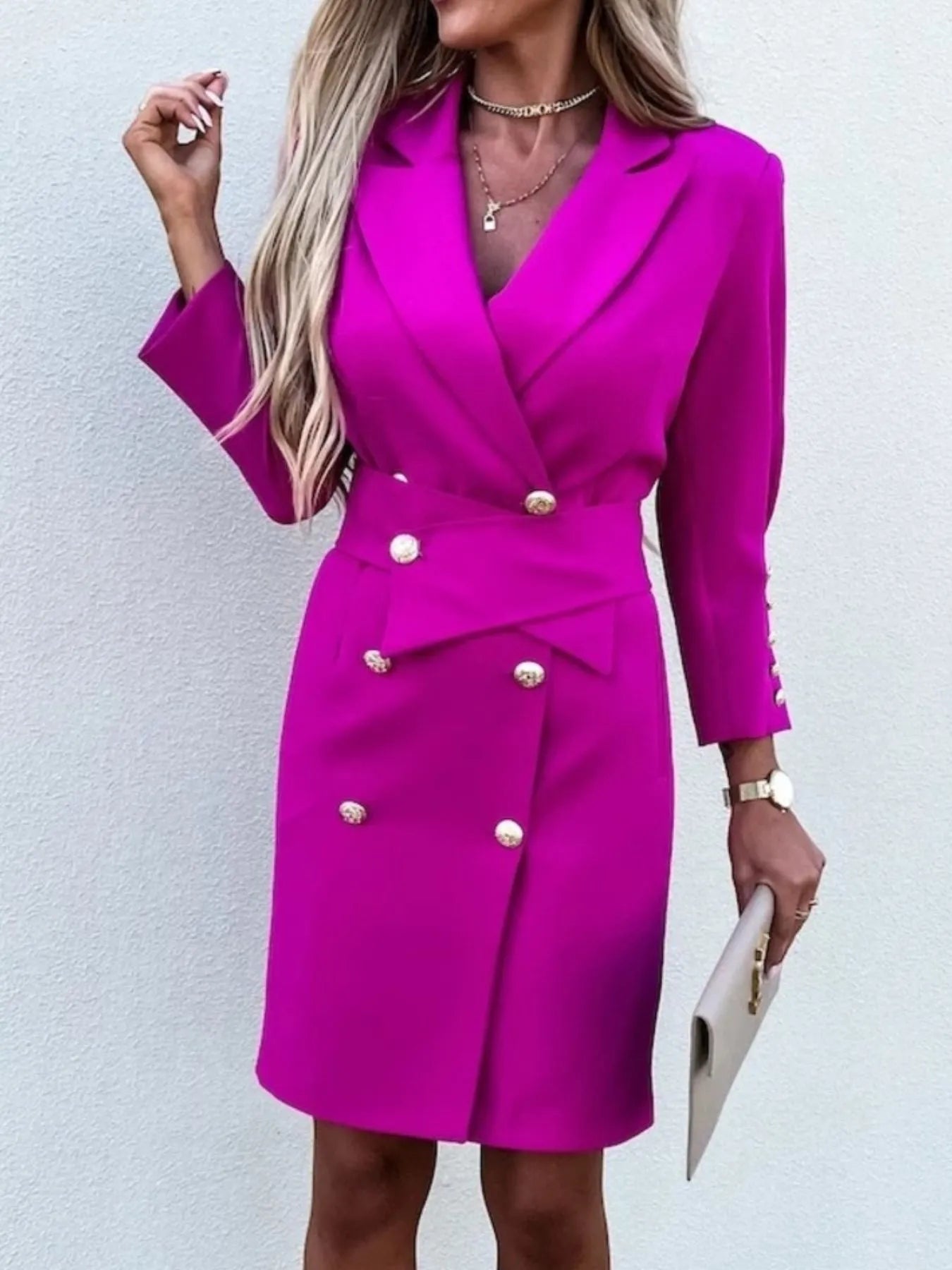 A woman in a stylish pink double-breasted blazer dress with gold buttons, accessorized with jewelry and a clutch.