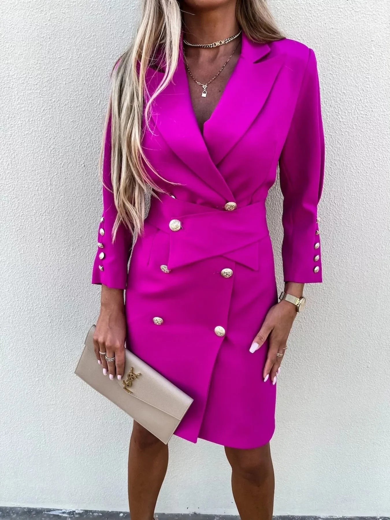 A woman in a stylish pink double-breasted blazer dress with gold buttons, accessorized with jewelry and a clutch.