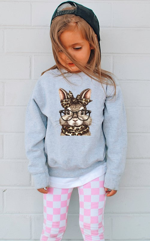 Child wearing a light blue sweatshirt with a cat graphic and pink checkered pants against a white brick wall.