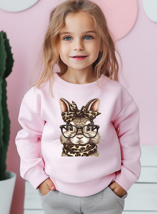 Child wearing a pink sweatshirt with a cat graphic, standing against a pink wall.