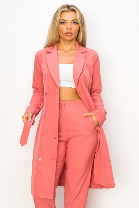 A woman rocking a salmon-colored trench coat with a belt and matching pants. She's got long blonde hair and her hands are casually in the coat's pockets.
