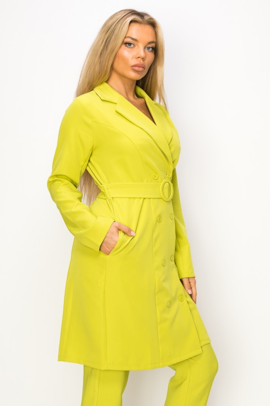 A woman wearing a bright yellow-green coat with a belt and matching pants. She's standing against a white background with her hands in the coat's pockets.