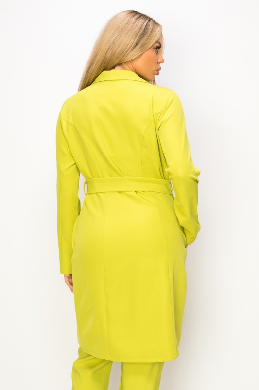 A woman wearing a bright yellow-green coat with a belt and matching pants. She's standing against a white background with her hands in the coat's pockets.