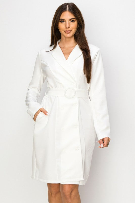 A woman wearing a crisp white belted coat dress with long sleeves and a collar. She's posing with one hand in her pocket and the other by her side, against a plain white background.