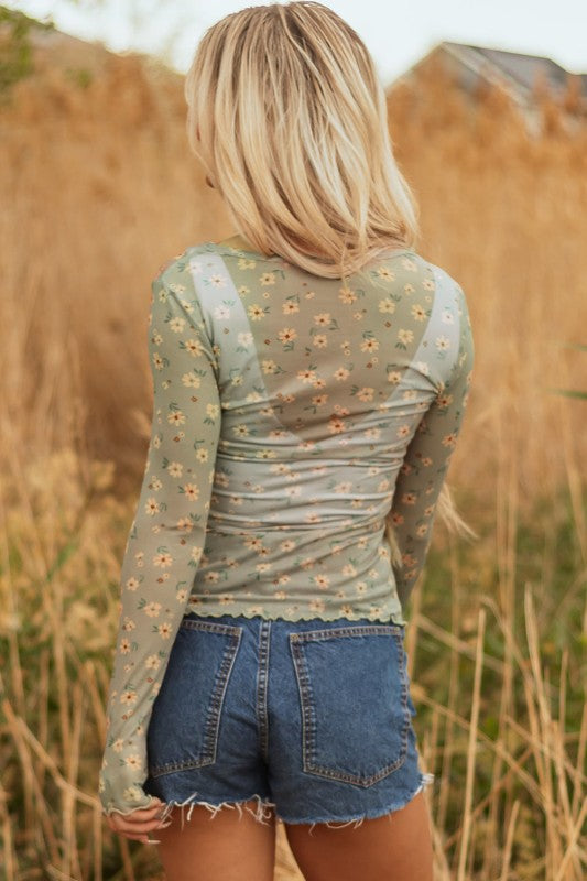 Lady in a sheer green floral top and frayed denim shorts, holding a straw hat, effortless, breezy style perfect for sunny outdoor days.