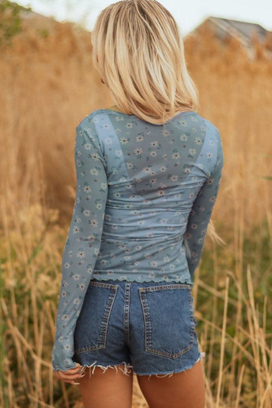 Lady in a sheer green floral top and frayed denim shorts, breezy style perfect for sunny outdoor days.