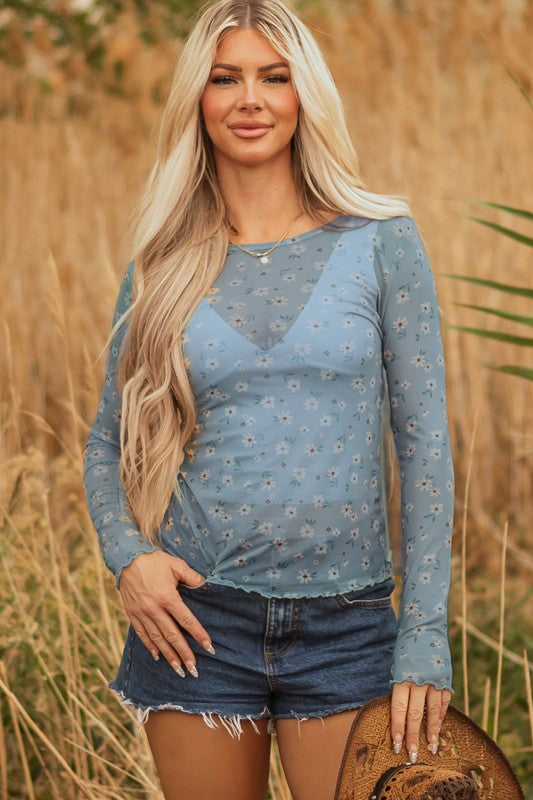 Lady in a sheer blue floral top and frayed denim shorts, holding a straw hat, effortless, breezy style perfect for sunny outdoor days.