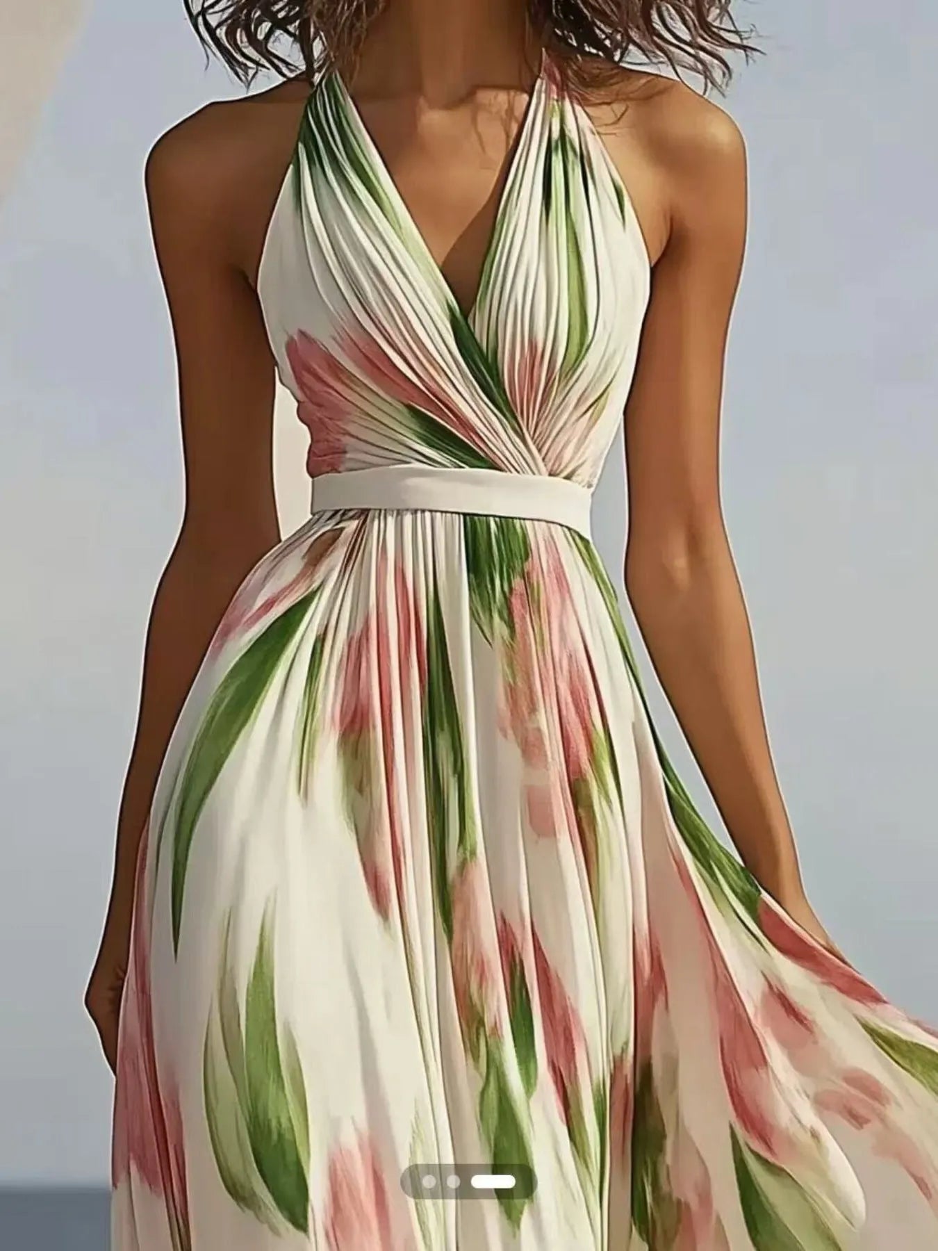 A woman wearing a flowy, white maxi dress with pink flowers and green leaves. It's a halter neck dress with a V-neckline and a belt at the waist. She's standing on a beach with the ocean behind her.