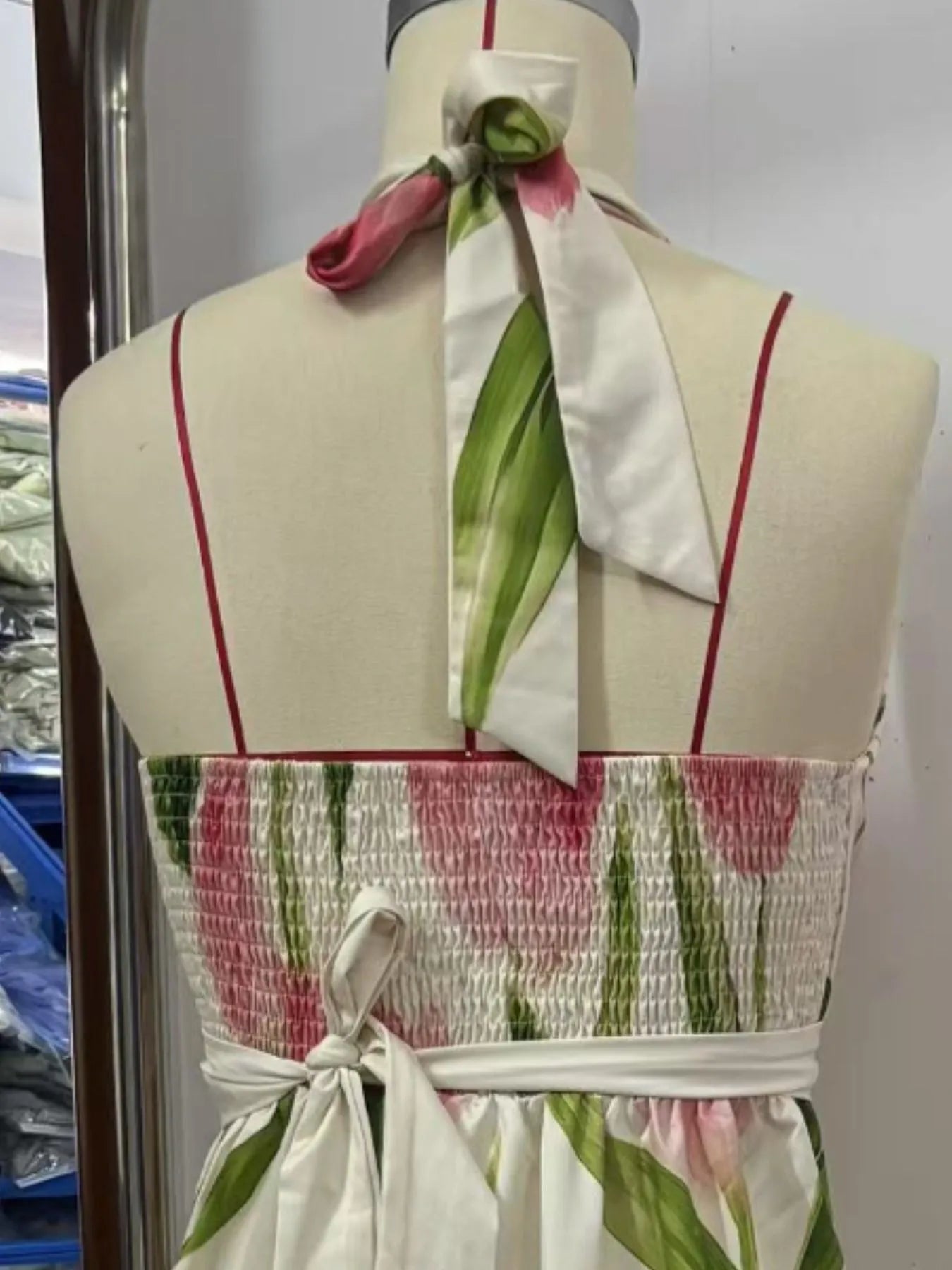 A flowy, white maxi dress with pink flowers and green leaves. It's a halter neck dress with a V-neckline and a belt at the waist.