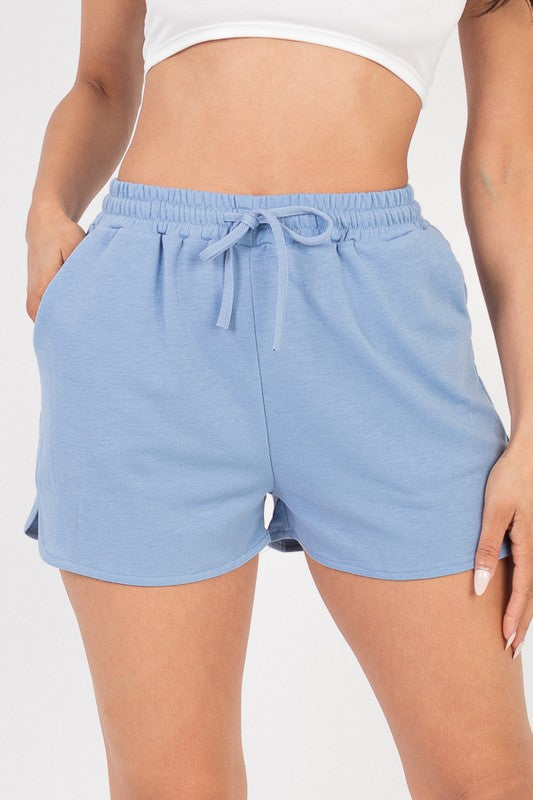 The image shows a woman wearing a white crop top and a blue shorts. She's posing with one hand in her pocket and the other on her thigh, against a plain white background.