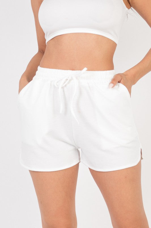 A woman wearing a white crop top and light white shorts with a drawstring waist, posing with one hand in her pocket against a plain background.